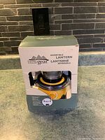 Lantern partially visible in worn open box on countertop showing yellow body and clear globe.