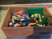 Box filled with assorted Lego pieces of different colors and sizes with some green baseplates visible.