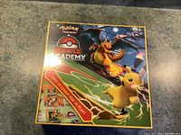 Front view of sealed Pokémon Battle Academy box with Pikachu and Charizard artwork shown on top of carpeted surface.