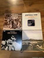 Lot of four vinyl records placed together on wooden floor, showing covers of Alice Cooper Greatest Hits, Johnny Cash Greatest Golden Hits Vol II, Elvis Presley On Stage February 1970, and The Moody Blues album.