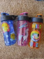 Photo shows three 16oz flip-top drinking cups with colorful designs on carpet background.