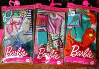Three Barbie accessory and outfit sets in original sealed pink plastic packaging with hangers displayed side by side.