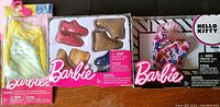 Photo showing three boxed sets of Barbie doll accessories including apparel and shoes. One set is yellow and white striped dress, another set is shoes in pink and gold colors, and a third set features Hello Kitty themed clothing.