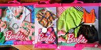 Photo showing three Barbie outfits in sealed plastic packaging with pink hangers, including flamingo print dress outfit, pumpkin sweater outfit, and neon green hoodie with tutu skirt outfit