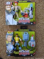 Photo of two Fisher Price Rescue Heroes action figures Billy Blazes and Carlos Kitbash in original packaging, showing character artwork, accessories, and branding.