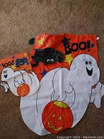 Photo showing two Halloween flags: one larger house flag with ghost holding pumpkin, black spider, and 'BOO!' text; one smaller garden/mailbox flag with ghost holding pumpkin and 'BOO!' text, all on orange backgrounds.