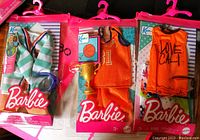 Photo showing three Ken Barbie male doll outfits in original pink packaging, including striped jacket, basketball uniform with trophy, and orange tank top outfit.