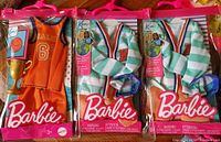Image showing three Ken doll male outfit accessory sets in their packaging: one orange basketball jersey outfit with trophy, two identical varsity jacket outfits with matching pants and belt, all branded Barbie and Mattel, new condition.