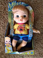 Photo of Littles Baby Alive Doll in original box, showing doll with blonde hair, yellow shirt with ladybug, and blue shorts.