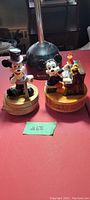 Two vintage Mickey Mouse music boxes displayed side by side on a red surface, one with Mickey dressed formally and the other at a piano.