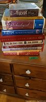 Stack of 9 hardcover books with one paperback in plastic on top, titles mostly about Egypt and some about China and Dan Brown novels.