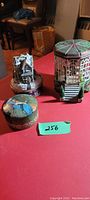 Three collectible music and trinket boxes shown together on red surface: a Norman Rockwell round box, Thomas Kinkade holiday gazebo-shaped box, and R & H Org Argyle & Fox music box with ship figure.