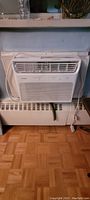 Front view of the white Noma air conditioner unit installed in a window with visible front vent and control panel.
