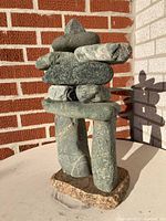 Photo shows a stone Inukshuk sculpture made from various grey-green stones stacked to form a figure, standing 18 inches tall in front of a brick and stucco wall.