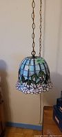 Full hanging view of Tiffany style stained glass lamp on chain over wooden floor and side table