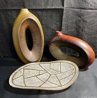 Two oval Artura ceramic vases in mustard yellow and reddish brown earth tones placed behind a footed geometric patterned clay plate.