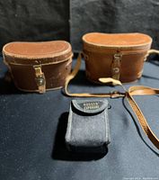 Two brown leather binocular cases and one black fabric case labeled Rugged Exposure shown on black surface.