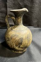 Front side view of the small Greek-style ceramic pitcher showing detailed human profile black-figure artwork on beige background with patina.