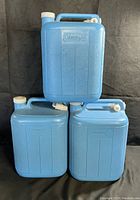 Three blue Coleman 5-gallon plastic water jugs stacked with visible handles and caps, two have dispensing spouts.