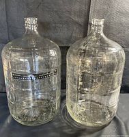 Front view of two clear glass carboy bottles side by side showing overall shape and condition with some residue on surfaces.