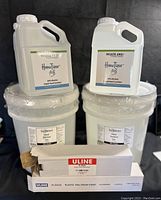 4 one gallon containers of liquid hand sanitizer labeled HanoTizer on top of 2 five gallon buckets of hand sanitizer gel, with 2 white pump dispenser boxes in front.