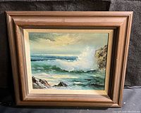 Front view of framed oil painting showing ocean wave, rocks, and sky.