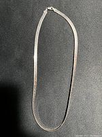 Full view of the silver necklace laid out on black background showing its flat, herringbone or snake chain style.