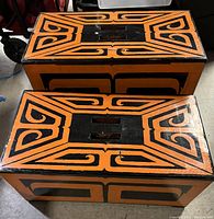 Top-down view of two rectangular art deco style side tables, showing black and orange geometric lacquered designs and the recessed carrying handles on top.
