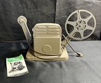 Vintage Baia Challenger film editor with crank handle, reel spindle, electrical cord and plug, sitting on a black background. Original user pamphlet shown beside.