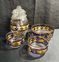 Full view of three bowls and one jar with lid, showing hand painted geometric bands in gold, blue and maroon on clear glass with craquelure and bubbles.