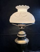 Full view of vintage brass lamp with milk glass shade showing eagle embossing on shade and scalloped top edge, lamp on black backdrop.