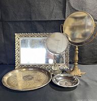 Full lot including brass pedestal mirror, chrome vanity mirror, filigree rectangular mirror, and three silver-colored trays of different sizes with engraved floral designs.