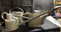 Photo showing two galvanized steel watering cans with curved handles and a galvanized bucket in the background on black backdrop.