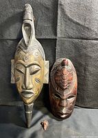 Three wooden carved masks including the tall Ivory Coast tribal mask, South Pacific turtle mask, and small talisman, all displayed on black fabric.