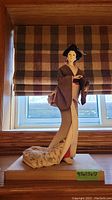 Full view of the porcelain Geisha statue standing on a wooden base by a window with a plaid curtain background.