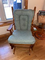 Front view of wooden glider rocker chair with padded, tufted greenish-blue cushion on seat and back, wooden spindle backrest visible.