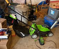 Lawn mower from side angle with grass collector bag, battery charger on floor, surrounded by garage items.