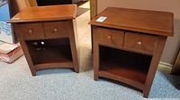 Pair of wood side tables showing front view with two drawers each and open storage cubby below. Surfaces show some wear.