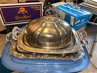 Set of four silver plated items displayed stacked; large dome lid, two trays, and ornate footed tray.