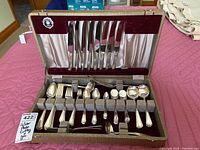 Open fibre board box with flatware shown. Box is lined with burgundy velvet and cream satin, with clips holding knives and flatware arranged in slots.