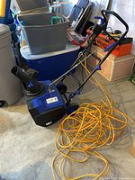 Side view of blue and black electric snow thrower with long orange extension cord on concrete floor, surrounded by storage bins and bags.