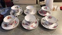 Top view of all six teacup and saucer sets arranged in two rows on countertop, showing various floral patterns and gold trimmed edges.