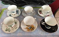 Photo showing six different teacups each with matching saucer, arranged in two rows. Various floral patterns and one black set, all with gold trimming.