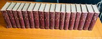 Full set of 18 hardcover books with burgundy faux leather spines and tan cloth covers, arranged side by side on a wooden surface.