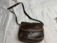 Front and top view of brown leather purse with flap closure and shoulder strap laid flat on white fabric.