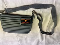 Front view of the dark gray ribbed rubber purse with adjustable gray fabric strap and metal clasps, showing the attached logo plaque.