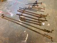 Overview photo showing 13 metal bar clamps arranged on the floor, longest clamp measuring approximately 77 inches, all clamps used with rust.