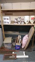 Overview of shelving unit containing several cans of paint, large glue container, white styrofoam piece, wooden air vents, cork board sheet, styrofoam chips in a bag, plastic bin and wood pieces, and a box of Clorox bleach