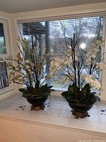 Two faux orchid flower arrangements displayed on a tiled windowsill with blinds in the background.