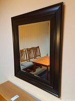 Full view of the rectangular mirror with dark wood frame hanging on the wall with dining chairs and table reflected in it.